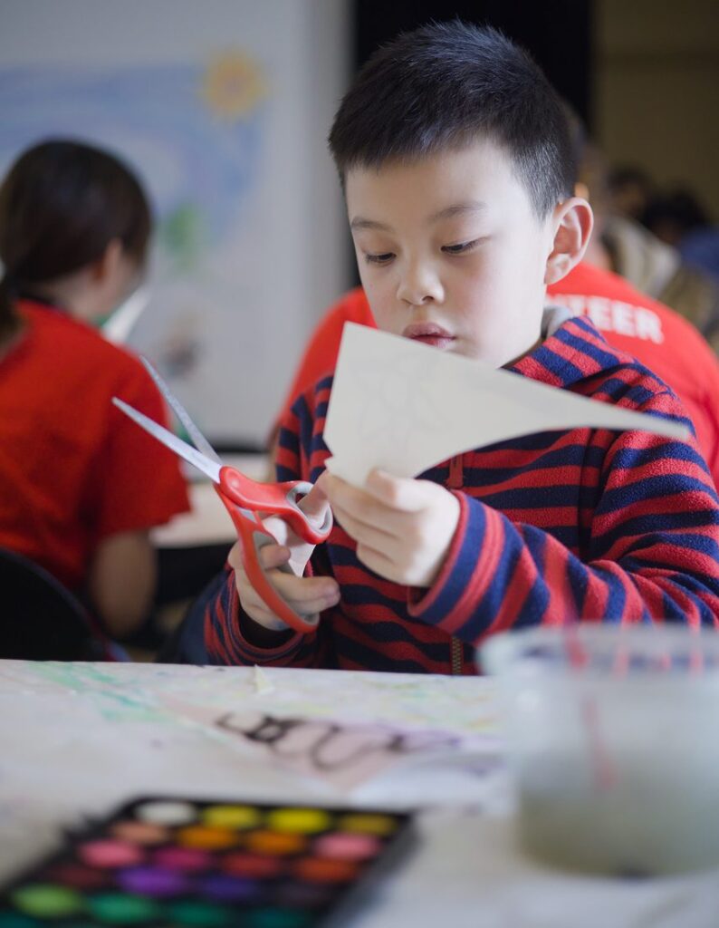 A young boy sits at a table with paints in front of him and cuts paper using red scissors.