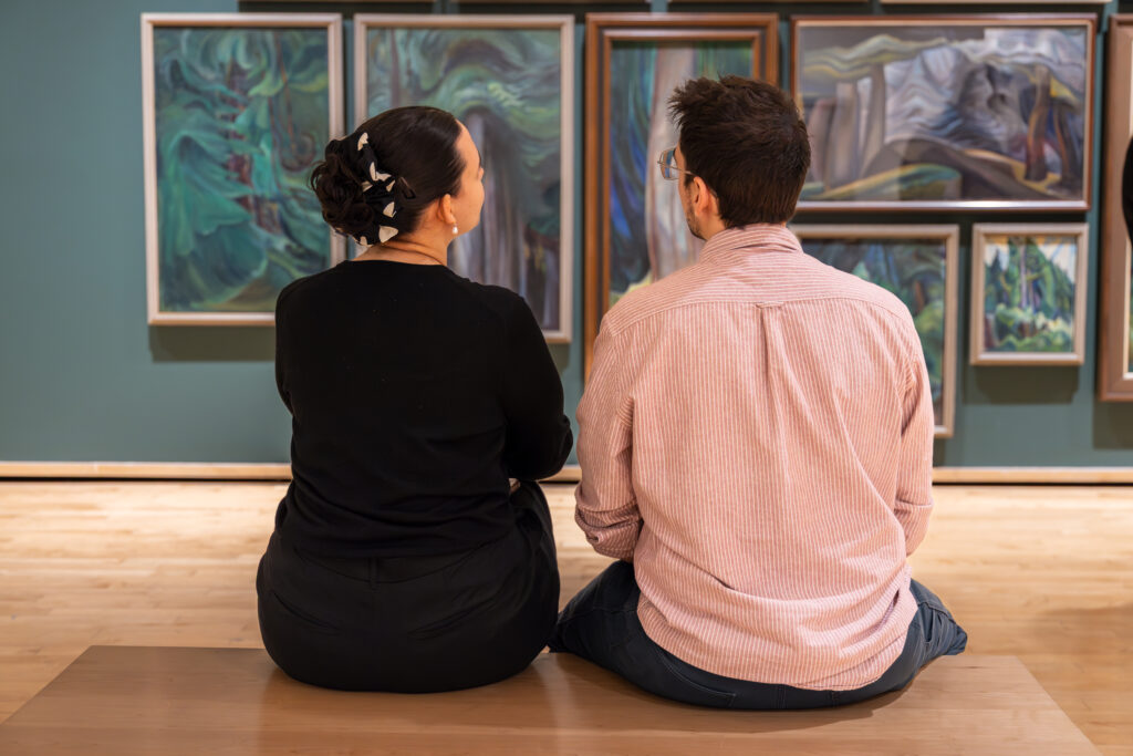 A woman and a man sit on a bench talking while looking at forest paintings by Emily Carr.