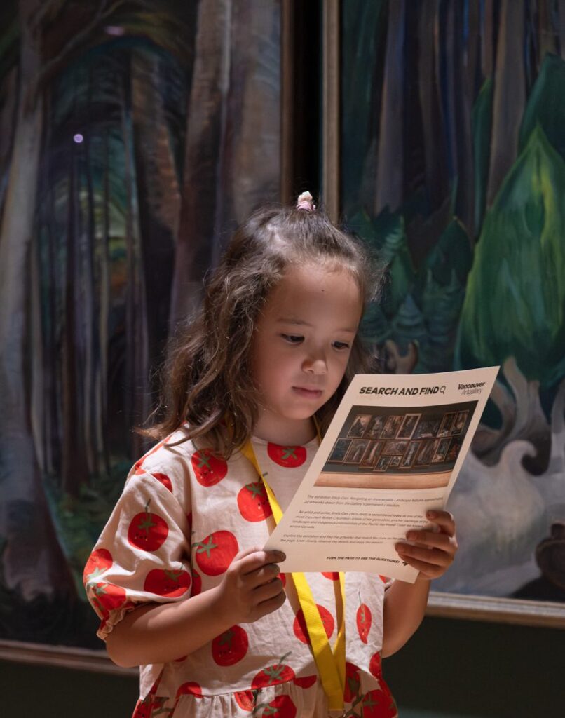 A young girl wearing a pink dress covered in a red tomato pattern stands in front of Emily Carr's forest paintings and holds the search-and-find activity in her hands.