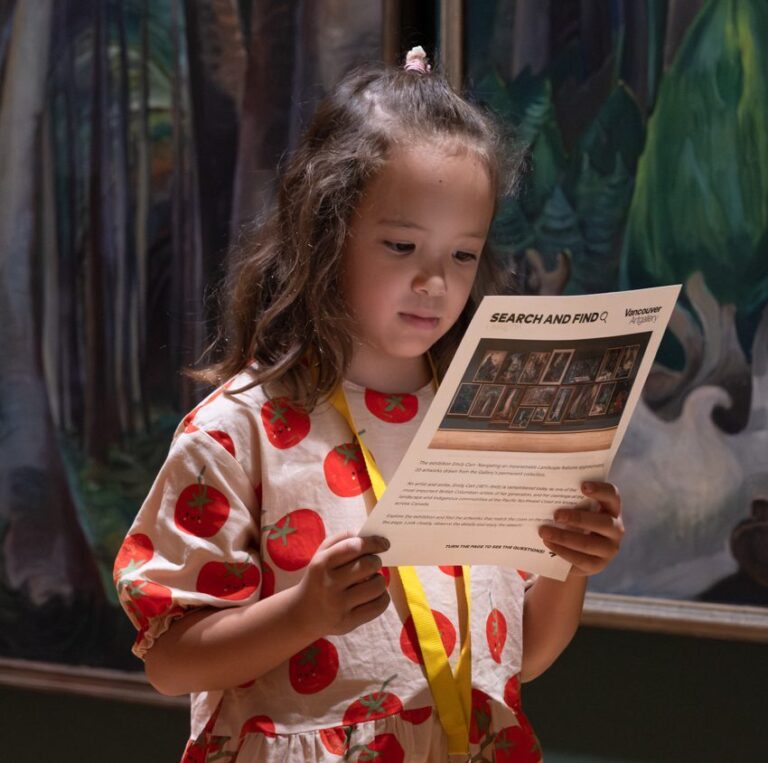 A young girl wearing a pink dress covered in a red tomato pattern stands in front of Emily Carr's forest paintings and holds the search-and-find activity in her hands.