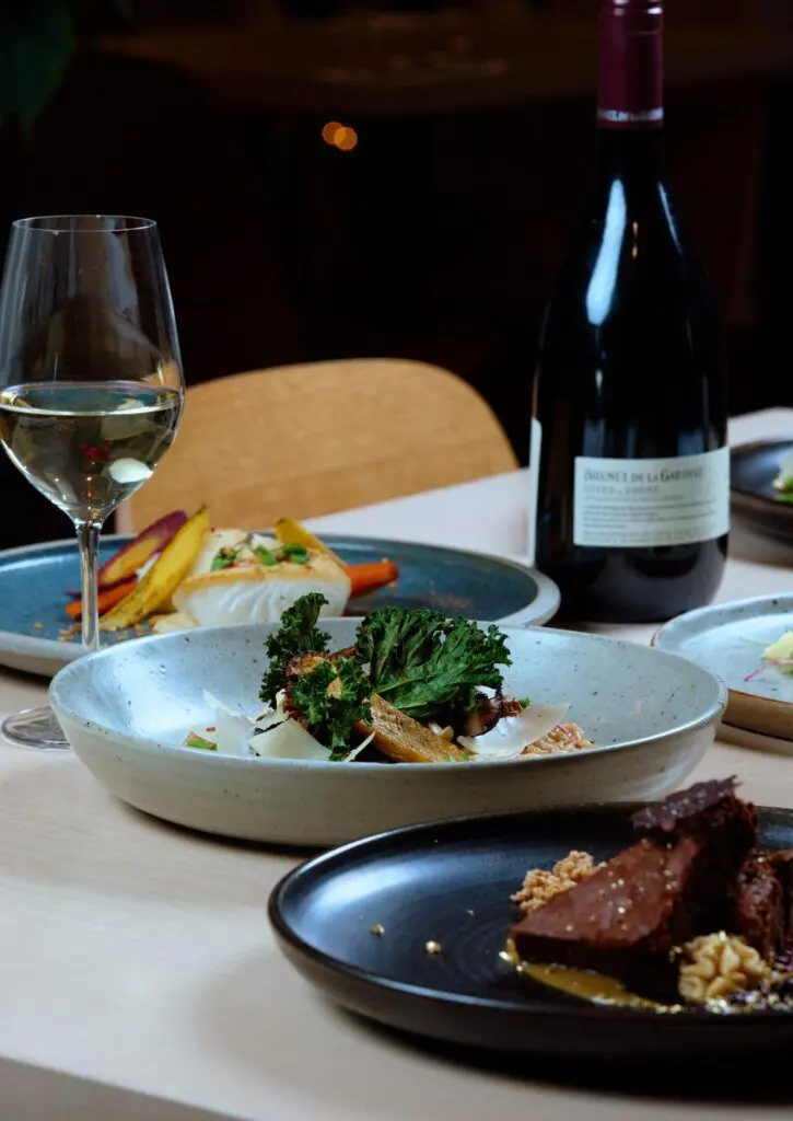 A table set with two dinner plates of meals from 1931 Gallery Bistro's 2025 Dine Out Vancouver menu, paired with a glass of wine and a wine bottle.
