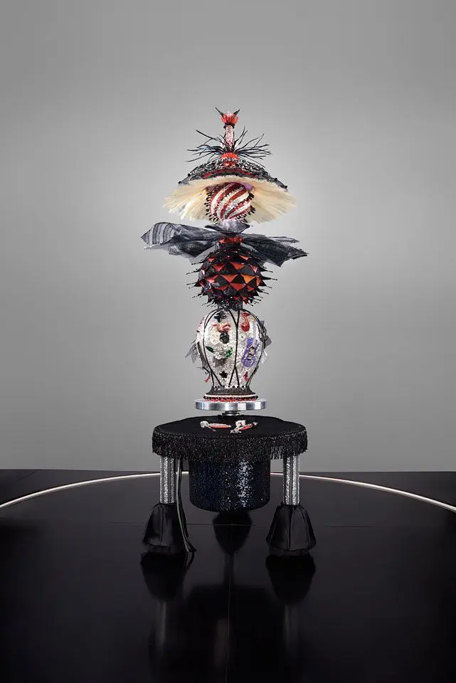 [Image Description: Centering’s shapely body is a stack of spheres and vase-like forms positioned atop a ceramic wheel. The sculpture is costumed in bedazzled textiles from hat-top to tripod bottom. Hand-applied and sewn beads, crystals, feathers, human hair, netting and sequins adorn its surface, suggesting a fantastical celebrity performance costume. On the flat velvet surface of the tripod, a collection of pottery tools has been similarly jeweled. Shown mid-spin, the costume splays outward by force of the swivel.]