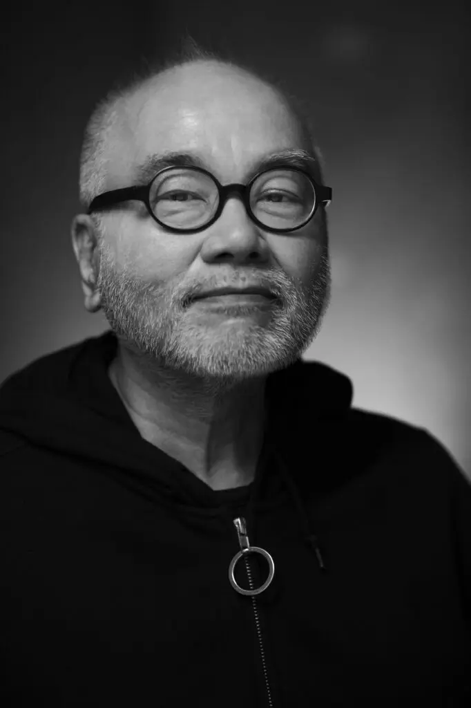 Black-and-white portrait of an Asian man with a short, white beard and circular glasses, seen from the chest up and wearing a black hoodie.