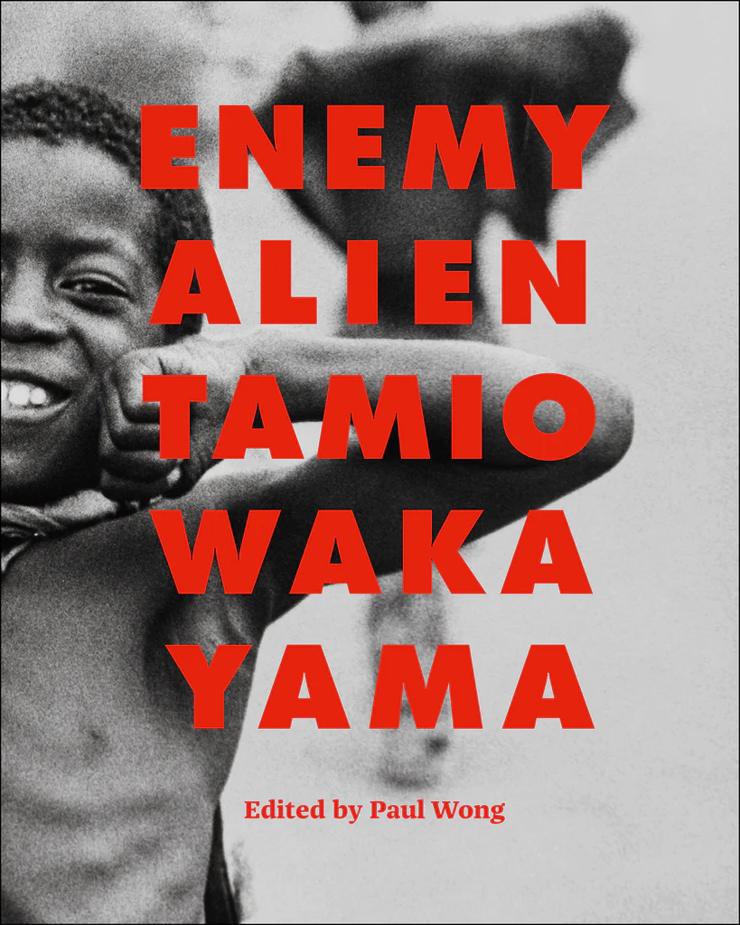 Cover of an exhibition catalogue with red, capital letters that read "Enemy Alien Tamio Wakayama." Below the title, in a smaller font, it reads: "Edited by Paul Wong." The red letters appear over a black-and-white photograph of a young, black boy with no shirt on, who smiles and beams out at the camera, while seemingly flexing his muscles.