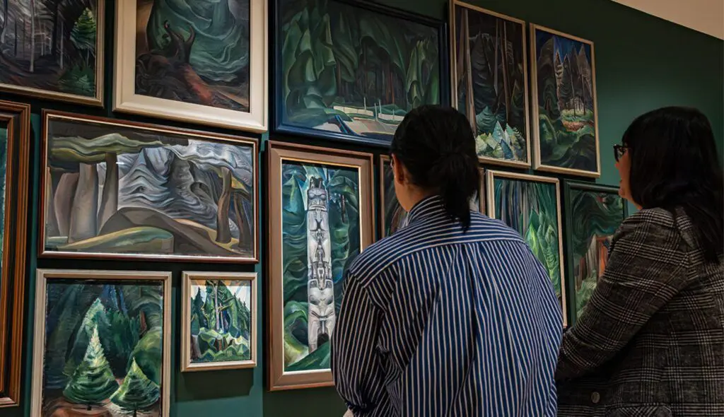 Two women seated looking at paintings of the forest by Emily Carr hung in a salon style.