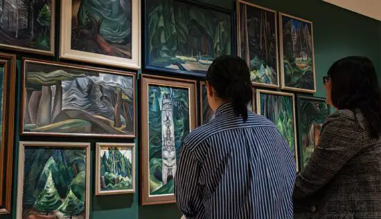 Two women seated looking at paintings of the forest by Emily Carr hung in a salon style.