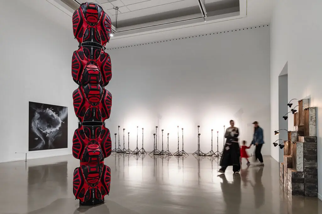 Installation view of the exhibition Postcards from the Heart: Selections from the Brigitte and Henning Freybe Collection. A sculpture that looks like a totem pole is visible in the foreground. It is made out of red golf bags. In the background, you can see an artwork of charcoal clouds, light stands and stacked boxes or cases. A family of three-a woman, man and young girl-enters the room. They are blurred.