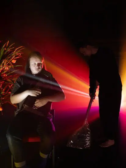 Two performers, one standing and another sitting, against a backdrop of red light.