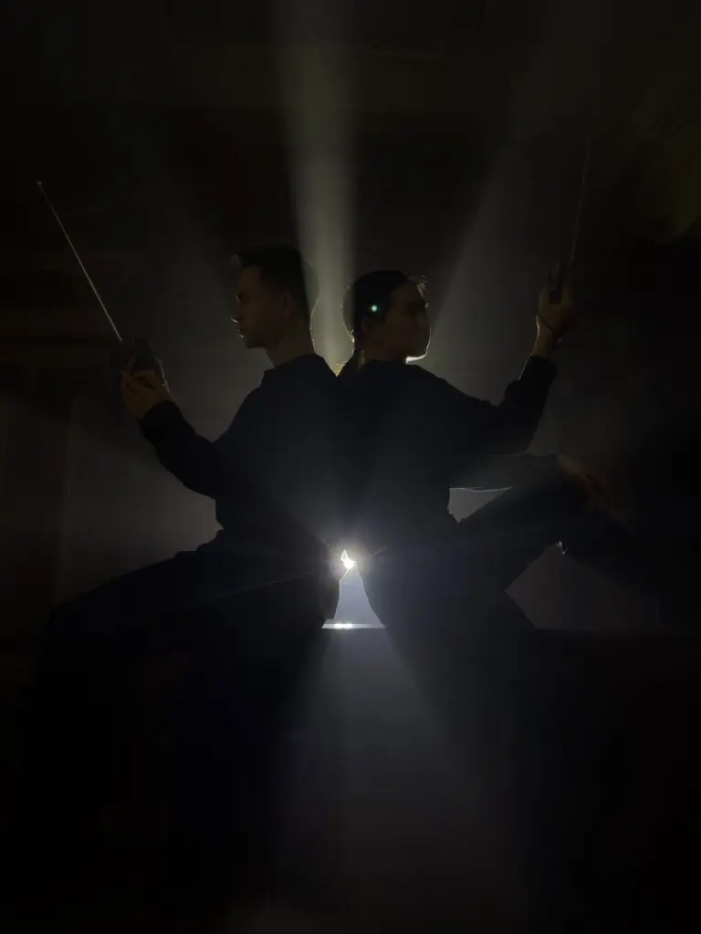 Two performers sitting back-to-back and holding handheld radios, in front of a spotlight.