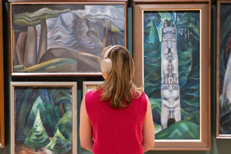 A visitor looking closely at paintings by Emily Carr while wearing headphones.