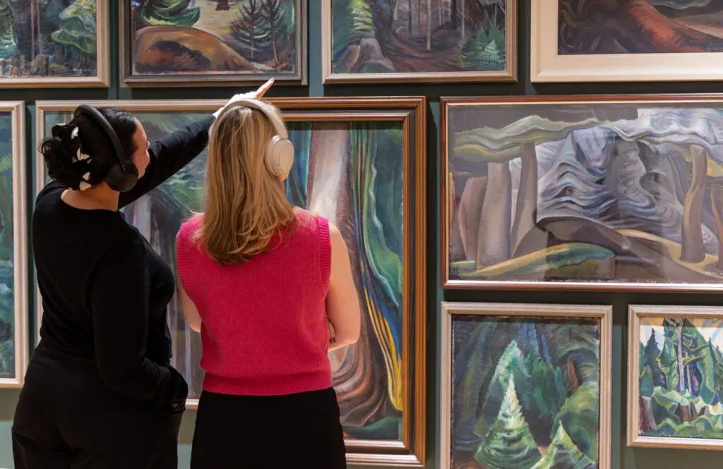 Two women stand and look at forest paintings by Emily Carr. One woman points at the paintings