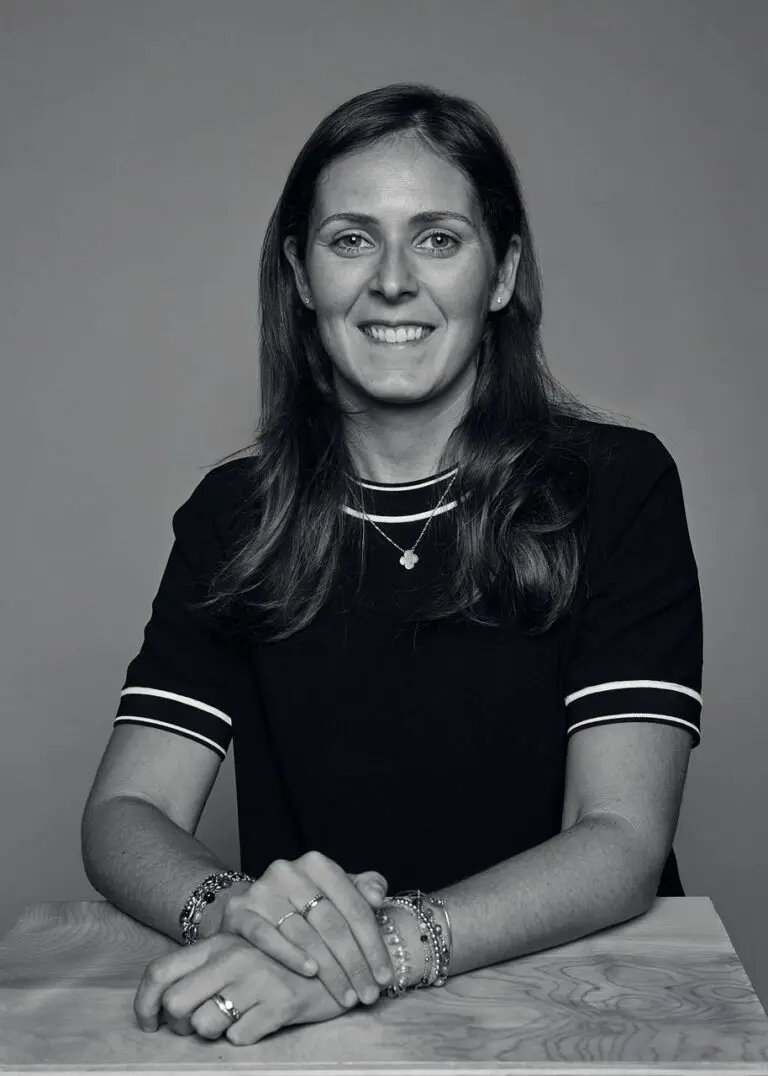 A black-and-white headshot of a woman from the waist up, with her hands place in front of her. She has light brown hair and olive skin tone and wears a black short sleeve top, a flower necklace and many bracelets.