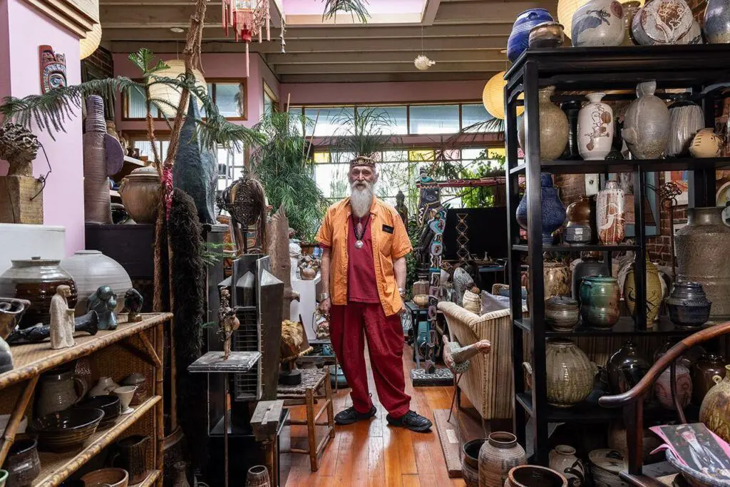 John David Lawrence, a man in his 70s with a white beard, stands in his home between shelves and shelves of ceramics.