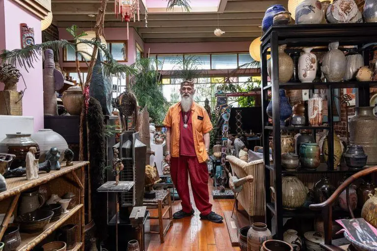 John David Lawrence, a man in his 70s with a white beard, stands in his home between shelves and shelves of ceramics.
