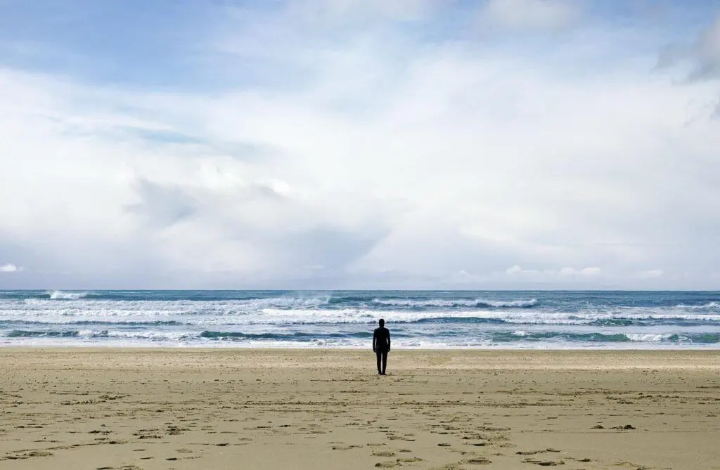 A beach scene with a single figure standing mid-frame on soft, beige sand that is textured with footprints. The figure looks out towards the wide-open ocean, which ripples with white-capped waves. The horizon is weighted by a blanket of dynamic clouds, and blue sky peaks out at the top of the frame.