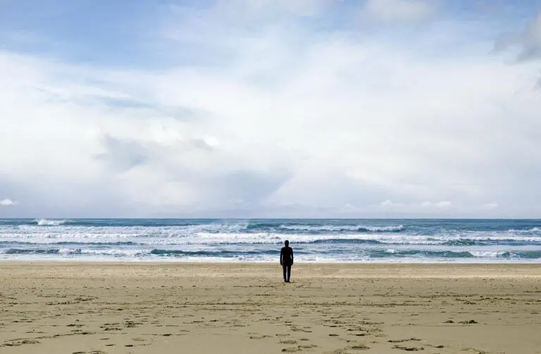 A beach scene with a single figure standing mid-frame on soft, beige sand that is textured with footprints. The figure looks out towards the wide-open ocean, which ripples with white-capped waves. The horizon is weighted by a blanket of dynamic clouds, and blue sky peaks out at the top of the frame.