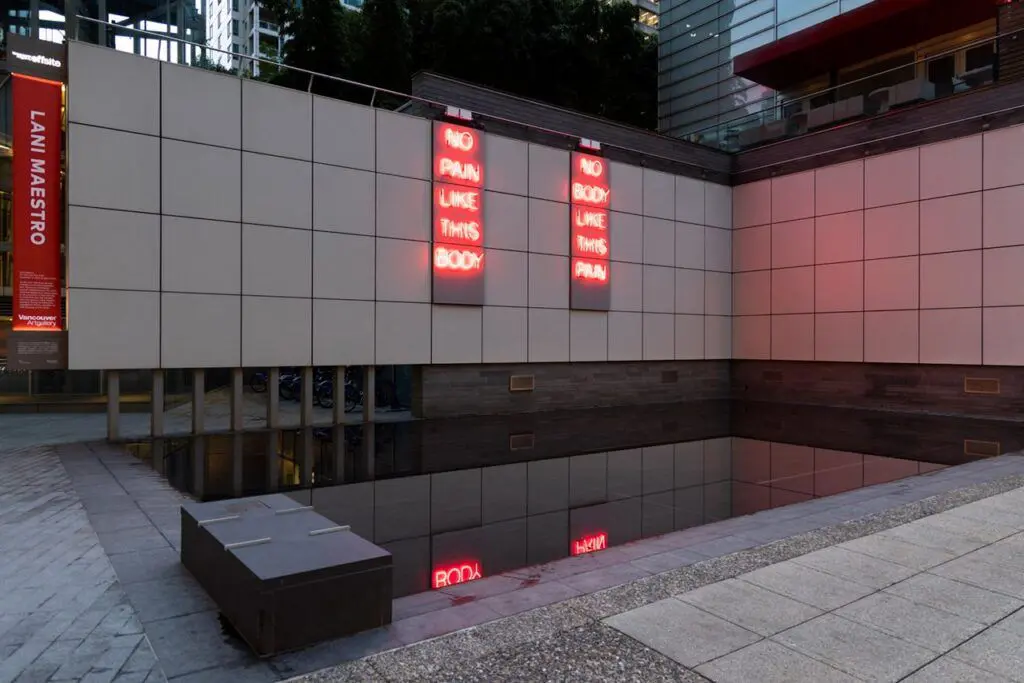 [Image Description: This work is a red neon sign diptych installed at the outdoor Vancouver Art Gallery Offsite at 1100 west Georgia Street. The exterior wall has a surface grid of large grey tiles. Each of the two neon statements fills three vertical tiles, installed two columns apart. The words are stacked vertically, one below the other. On the left, text reads: “No Pain Like This Body.” On the right, text reads: “No Body Like This Pain.”]