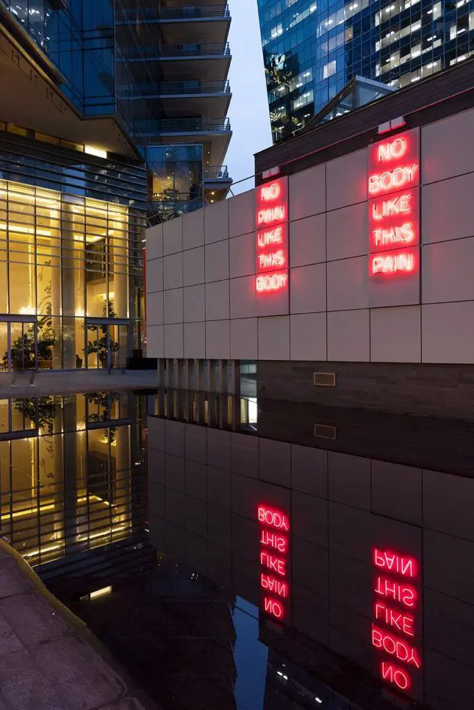 [Image Description: This work is a red neon sign diptych installed at the outdoor Vancouver Art Gallery Offsite at 1100 west Georgia Street. The exterior wall has a surface grid of large grey tiles. Each of the two neon statements fills three vertical tiles, installed two columns apart. The words are stacked vertically, one below the other. On the left, text reads: “No Pain Like This Body.” On the right, text reads: “No Body Like This Pain.”]