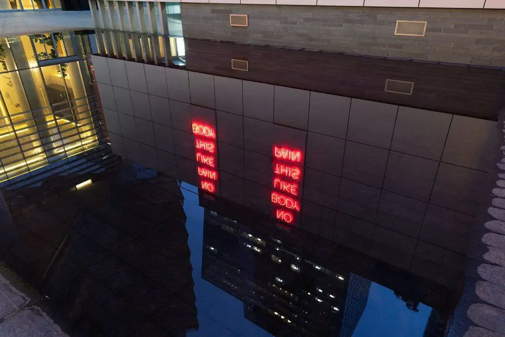 [Image Description: This work is a red neon sign diptych installed at the outdoor Vancouver Art Gallery Offsite at 1100 west Georgia Street. The exterior wall has a surface grid of large grey tiles. Each of the two neon statements fills three vertical tiles, installed two columns apart. The words are stacked vertically, one below the other. On the left, text reads: “No Pain Like This Body.” On the right, text reads: “No Body Like This Pain.”]