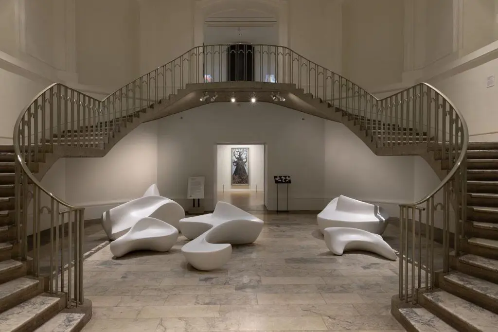 Five white, hand-carved curvilinear forms are installed on the rotunda’s marble floor, and framed by curving staircases on either side. The forms are large enough to sit on, with smooth, flat surfaces that pinch and peak into back or arm rests. In the background, a doorway into the gallery shows an oil painting of a tree.