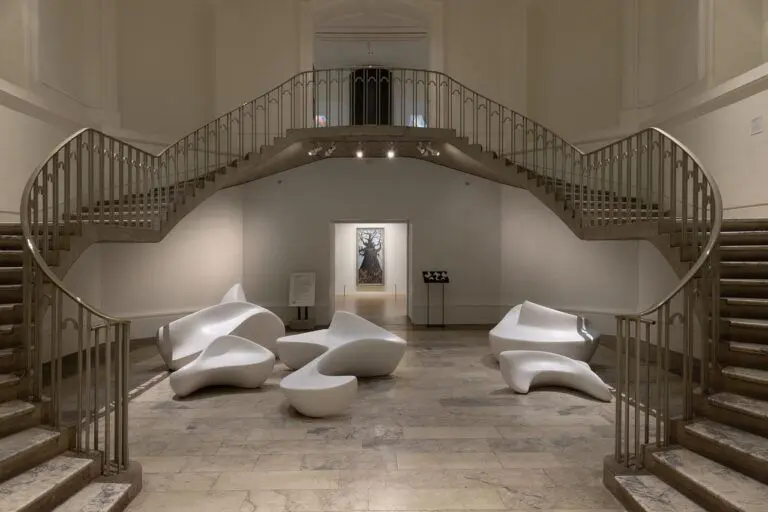 Five white, hand-carved curvilinear forms are installed on the rotunda’s marble floor, and framed by curving staircases on either side. The forms are large enough to sit on, with smooth, flat surfaces that pinch and peak into back or arm rests. In the background, a doorway into the gallery shows an oil painting of a tree.