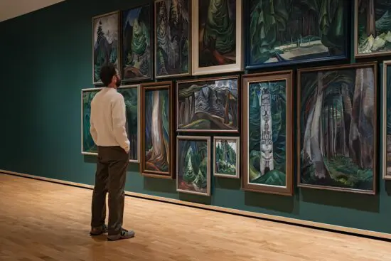 Man wearing a white sweater and black pants stands in front of a wall of Emily Carr paintings of forest landscapes. The wall is also painted a forest green.
