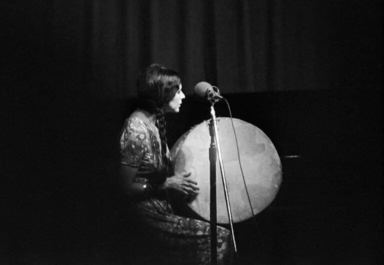[Image Description: A black and white photograph of Alanis Obomsawin on stage. She is seated and wearing a floor length floral-print dress. Her hair hangs in two long braids over her shoulders. She holds a large hand drum, her right hand ready to play. The texture on the drum’s surface is emphasized by the grain of the film; it glows like a full moon. A microphone is positioned in the foreground.]