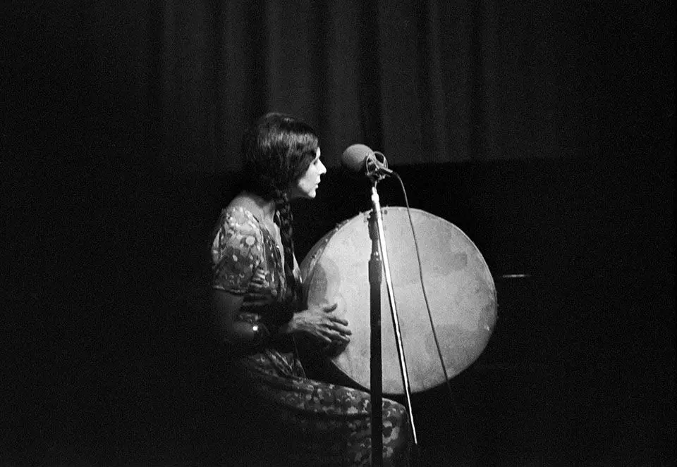 [Image Description: A black and white photograph of Alanis Obomsawin on stage. She is seated and wearing a floor length floral-print dress. Her hair hangs in two long braids over her shoulders. She holds a large hand drum, her right hand ready to play. The texture on the drum’s surface is emphasized by the grain of the film; it glows like a full moon. A microphone is positioned in the foreground.]