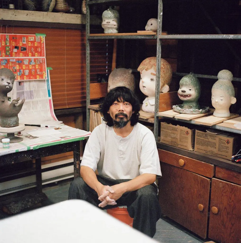 A man, Japanese artist Otani Workshop, sits in a studio and looks out at the camera. There are unfinished ceramic sculptures of a beaver, boy, dinosaur and woman behind him on metal and wooden shelving.