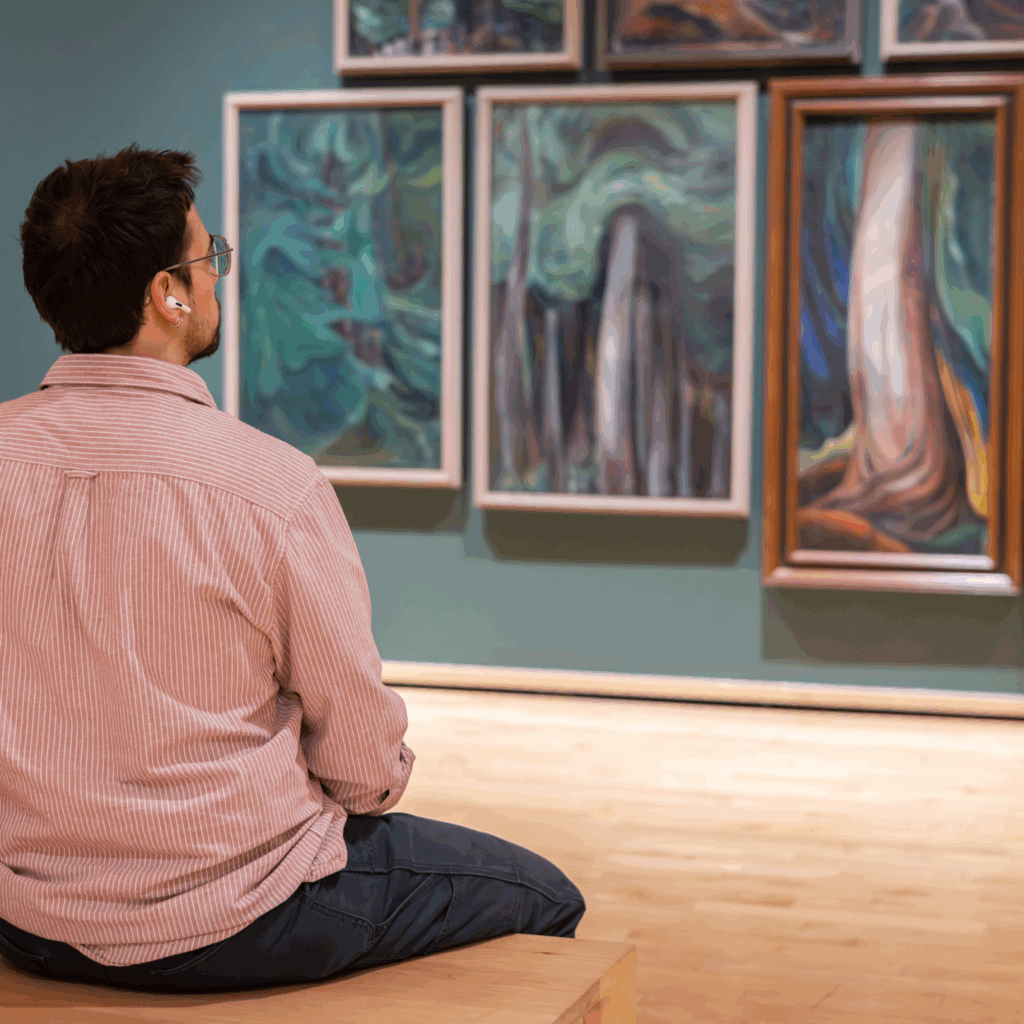 Man on a pink shirt sits on a bench and looks at forest paintings by Emily Carr