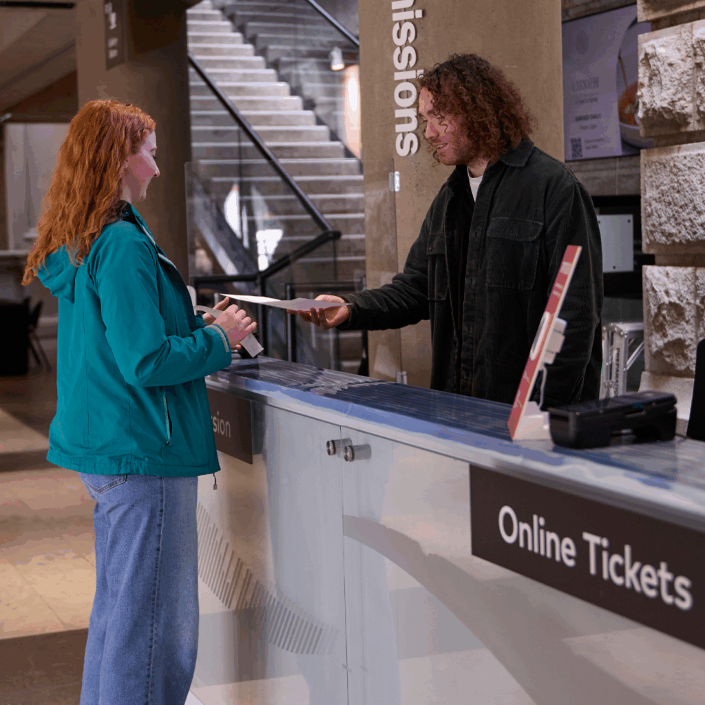 A woman with red hair in a green jacked checks in at the Admissions Desk and speaks to a man with curly blond hair. She receives a brochure from the man at the desk.