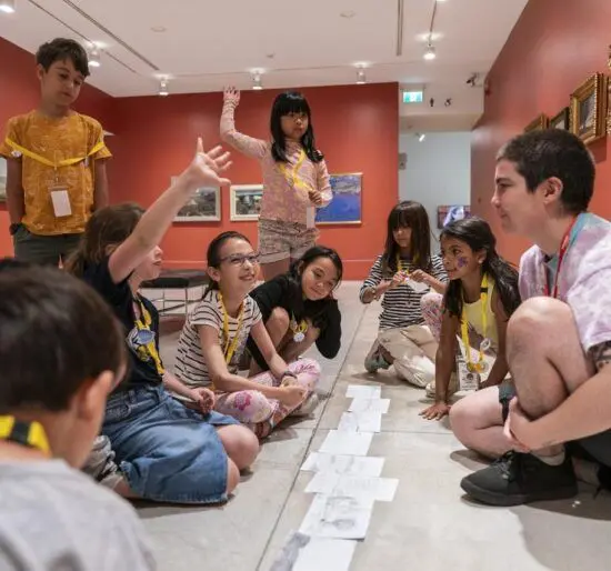 Children sit in the exhibition space in a circle with a camp leader. A child has their hand up.