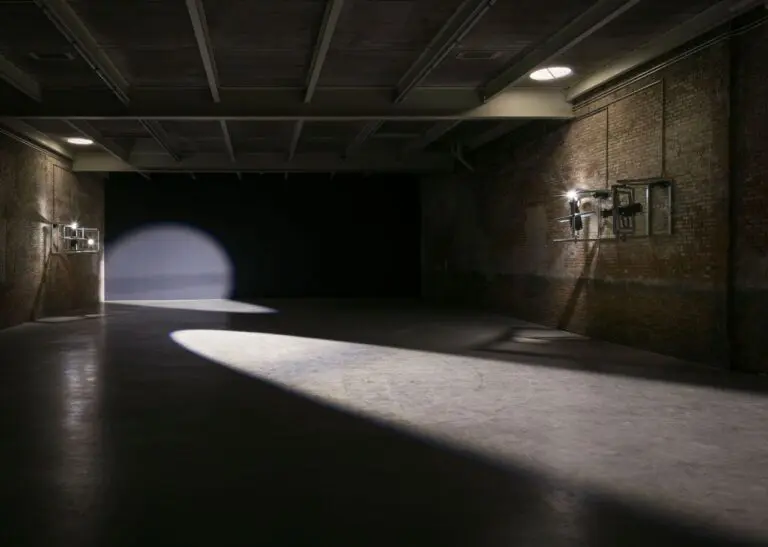 Installation view of Lucy Raven's Casters featuring lights mounted to the wall and creating a spotlight projecting a cleaved circle of light on the floor.