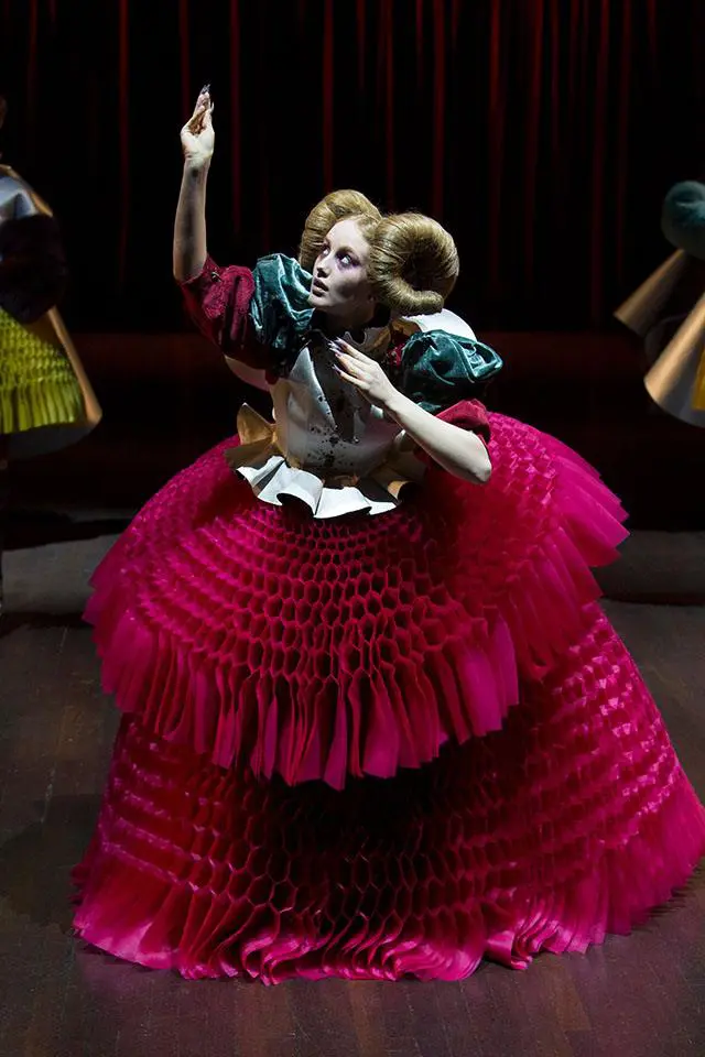 [Image Description: A model with powdery white skin is posed with arms held upwards like a marionette on a dark stage in front of a red velvet curtain. She is wearing a dress designed by the Japanese designer Jun Takahashi. The dramatic garment includes a form-fitted, corset style jacket with puffed sleeves and a voluptuous, fuchsia bustle skirt constructed of silk and nylon ribbons that are sewn into a honeycomb pattern. The model’s golden hair is tied up into large donut-shaped buns, one on either side of her head.]