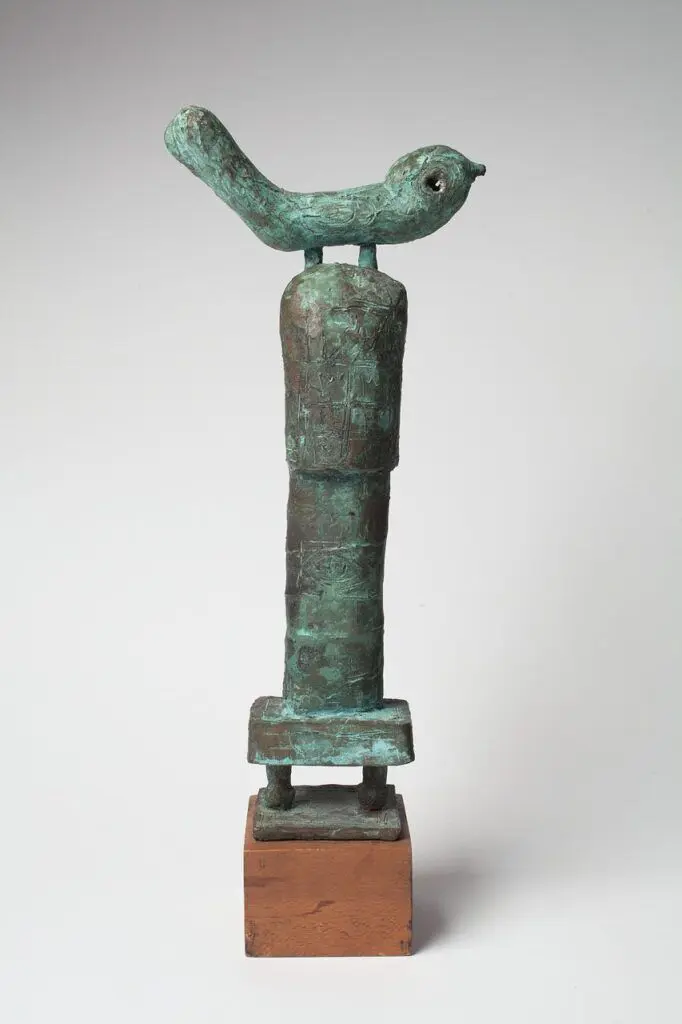 [Image Description: A studio shot of the sculpture positioned on a white backdrop: a bronze representation of a small bird, perched on top of a vertical pillar. The bronze is aged and oxidized, showing a pastel green patina. It is mounted onto a small wooden cube.]