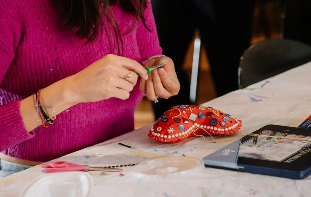 Detail of a woman's hands as she strings beads on a thread to sew onto a felt heart, sitting on the table in front of her.