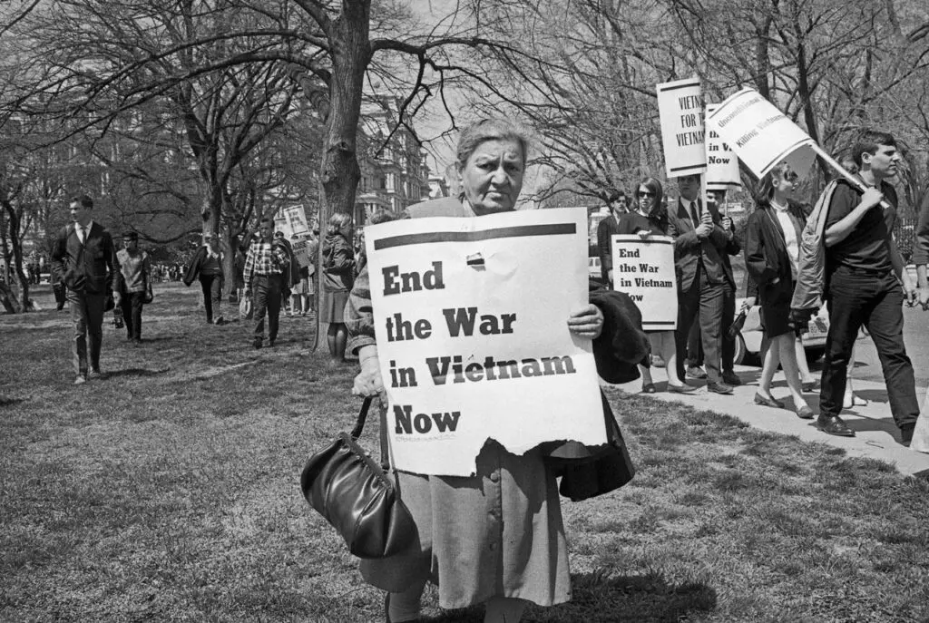 Black-and-white photograph of a woman holding a newspaper in a park amidst a protest. The cover of the newspaper says: End the War in Vietnam Now.