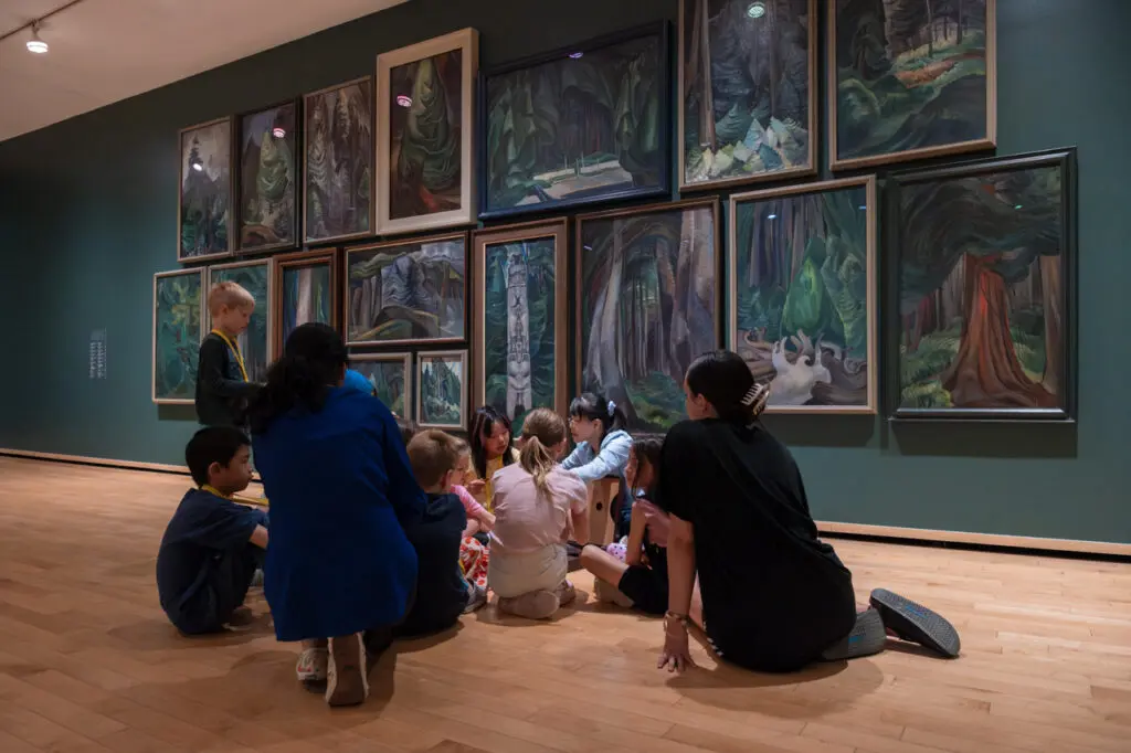 A group of children and adults sit in the Gallery in front of a wall of Emily Carr paintings of trees on a tour.
