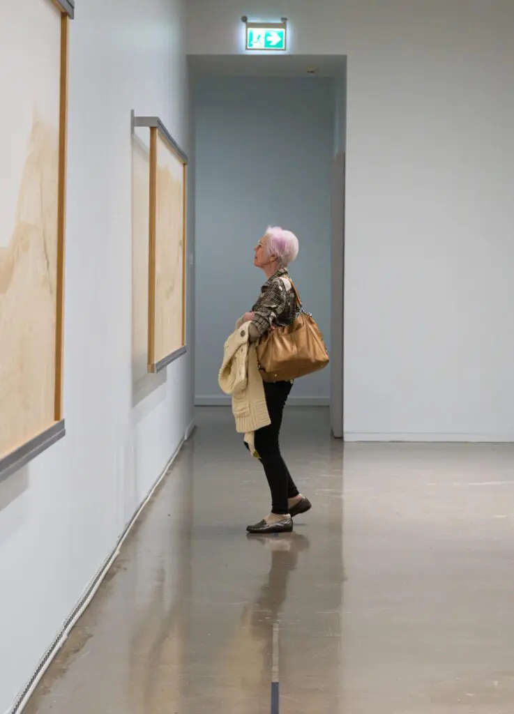 A woman with white hair and a pink highlight looking at works hanging on a wall