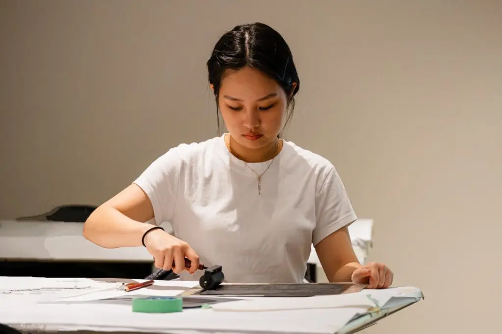 A young woman wearing a white shirt sits at a table and uses a roller to press a paper to make a print.