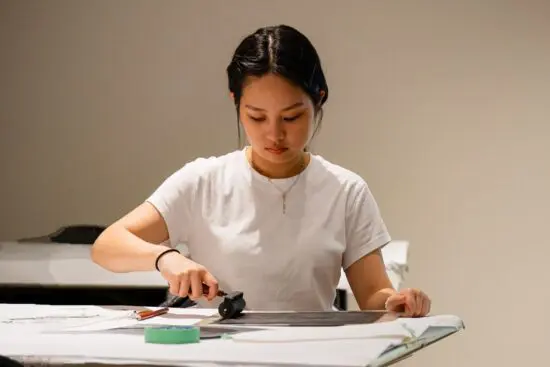 A young woman wearing a white shirt sits at a table and uses a roller to press a paper to make a print.