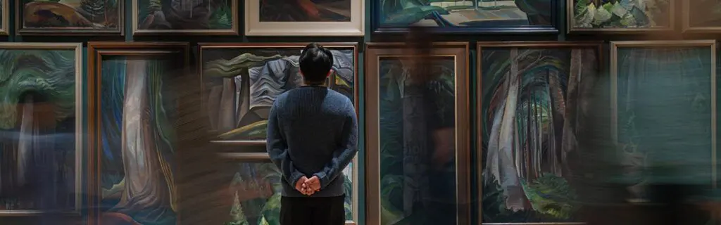 Man stands still, looking at Emily Carr paintings, while others rush by